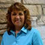 A photo of a woman with should length hair wearing a blue blouse Carol is smiling into the camera standing in front of a stone background