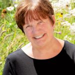 A woman with ear length red hair and blue eyes smiles into the camera. She is wearing a black blouse. The background is a field of flowers