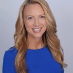 A woman with shoulder length blonde hair smiles at the camera. She is wearing a blue blouse and stands in front of a gray background